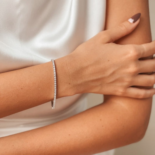 Nahaufnahme eines Frauenarms, der ein elegantes, silbernes Tennisarmband mit funkelnden Zirkonia-Steinen trägt, vor weißem Satin-Hintergrund.