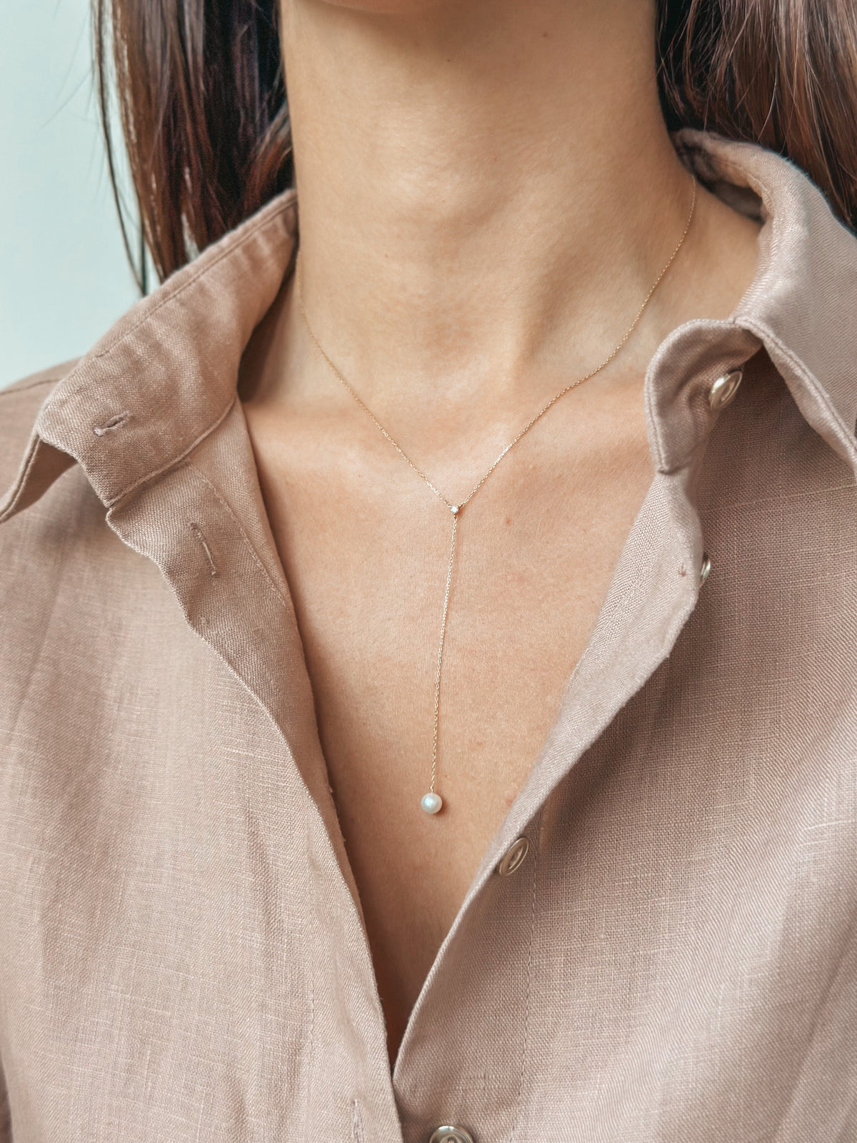 A woman wearing a gold necklace with a small pearl pendant, partially visible beige shirt