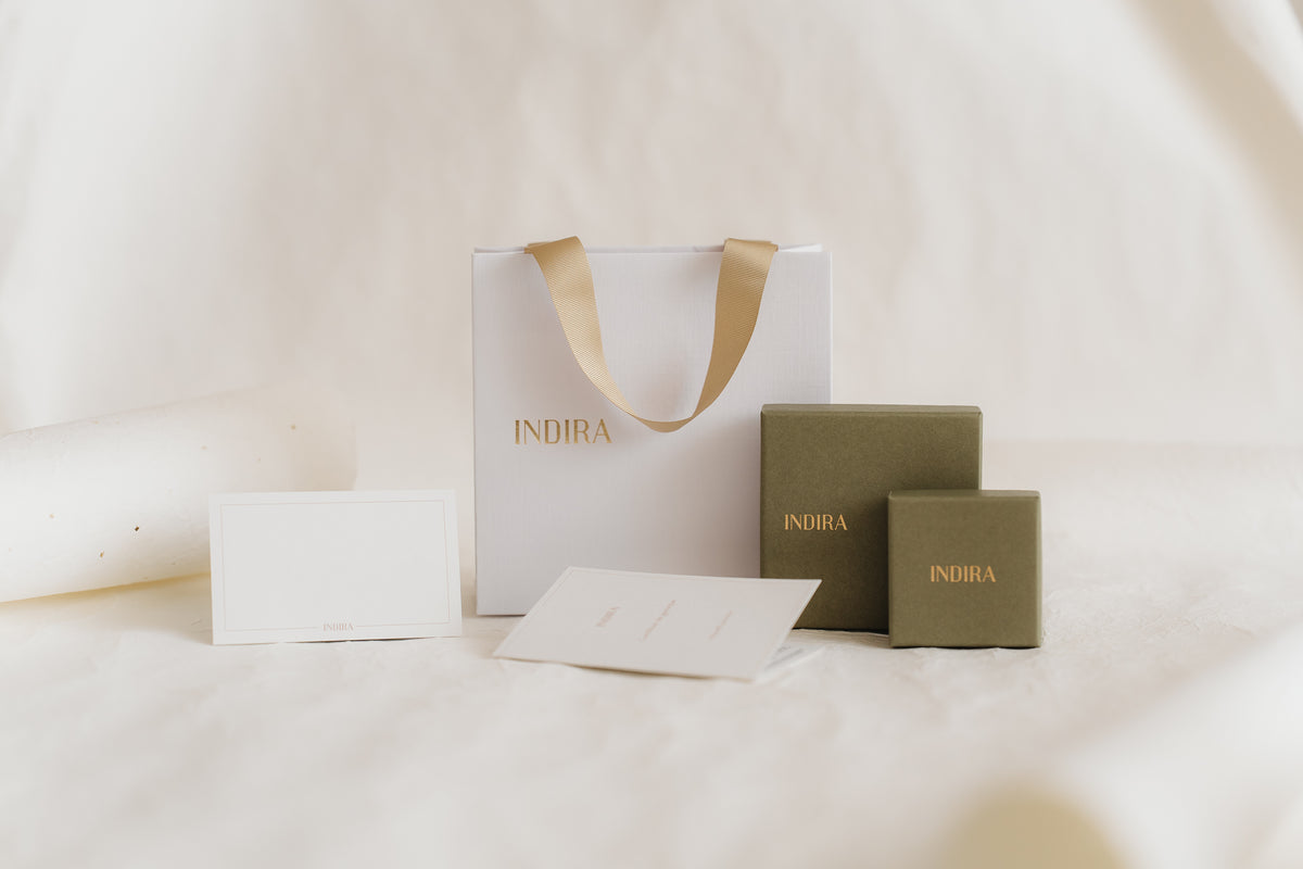 Minimales Studiofoto des INDIRA-Geschenkverpackungssets: eine weiße Geschenktüte mit Henkeln aus goldfarbenem Band, zwei olivgrüne Schmuckschachteln und leere Karten auf weichem elfenbeinfarbenem Hintergrund.
