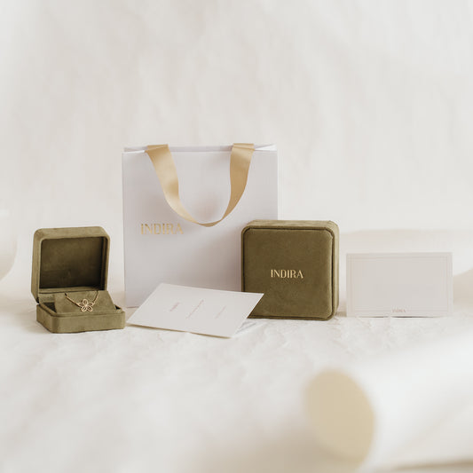 Indira Geschenkverpackungs-Set mit einer weißen Geschenktasche mit goldenen Bandhenkeln, olivfarbenen Samt-Schmuckschachteln (eine geöffnet) und Karten, arrangiert auf einem weichen, neutralen Hintergrund.