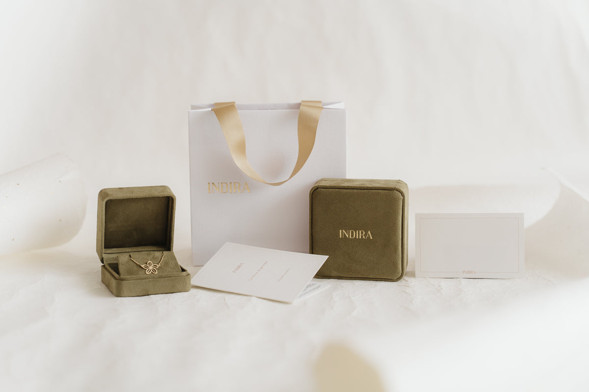 Indira Geschenkverpackungs-Set: weiße Geschenktasche mit goldenen Bandhenkeln, zwei olivgrüne Samt-Schmuckschatullen (eine geöffnet und zeigt einen goldenen Blumenanhänger) sowie Karten mit Markenlogo auf einem weichen, weißen Studiohintergrund.