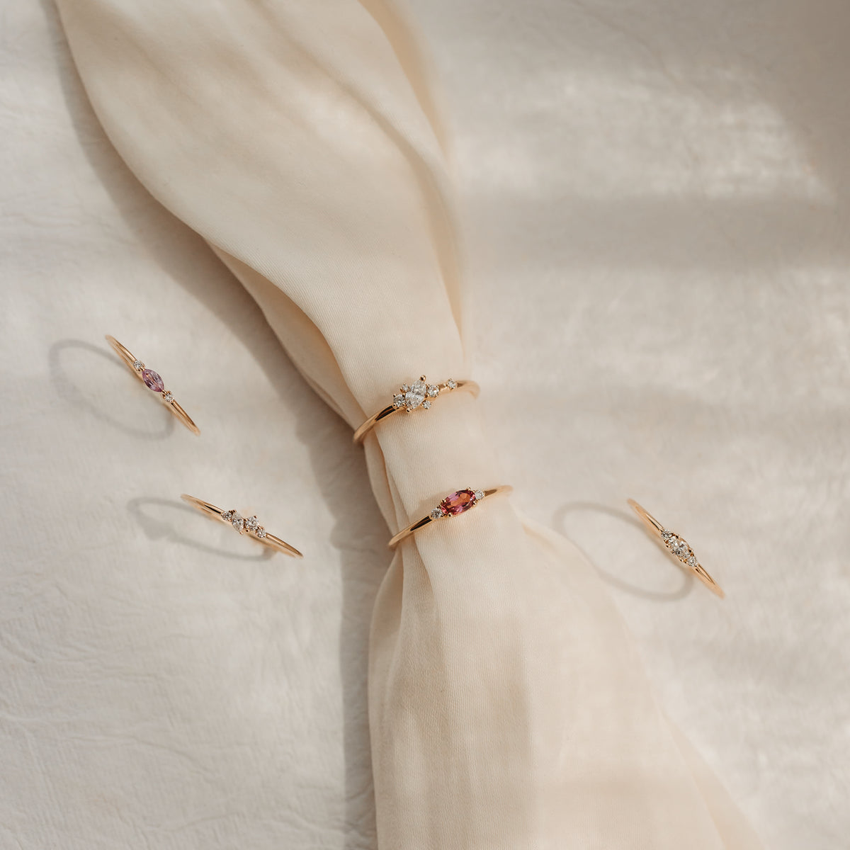 Fünf zarte 14k Gelbgold-Ringe mit Diamanten und rosafarbenen Edelsteinen, arrangiert auf geknotetem cremefarbenem Seidenstoff im sanften Sonnenlicht.