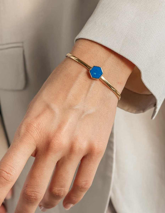 Nahaufnahme eines Handgelenks, das ein goldenes Armband mit einem blauen, sechseckigen Stein trägt.