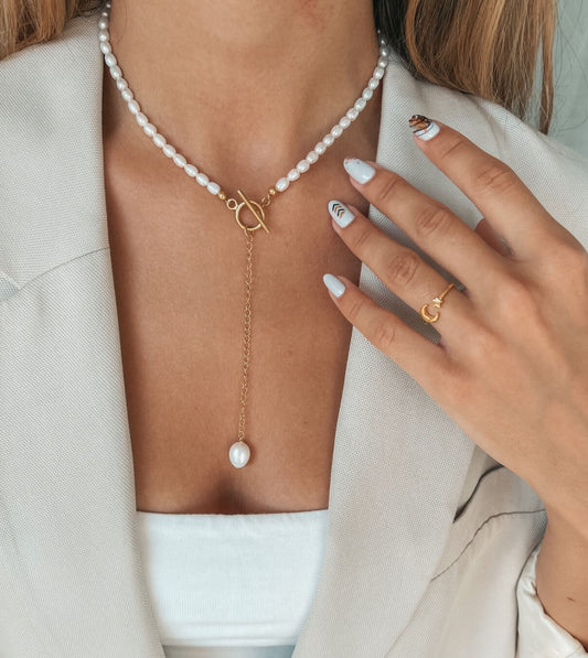 Nahaufnahme eines Frauenhalses mit einer goldenen Halskette und hängendem Perlenanhänger, ergänzt durch einen minimalistischen Goldring am Finger.