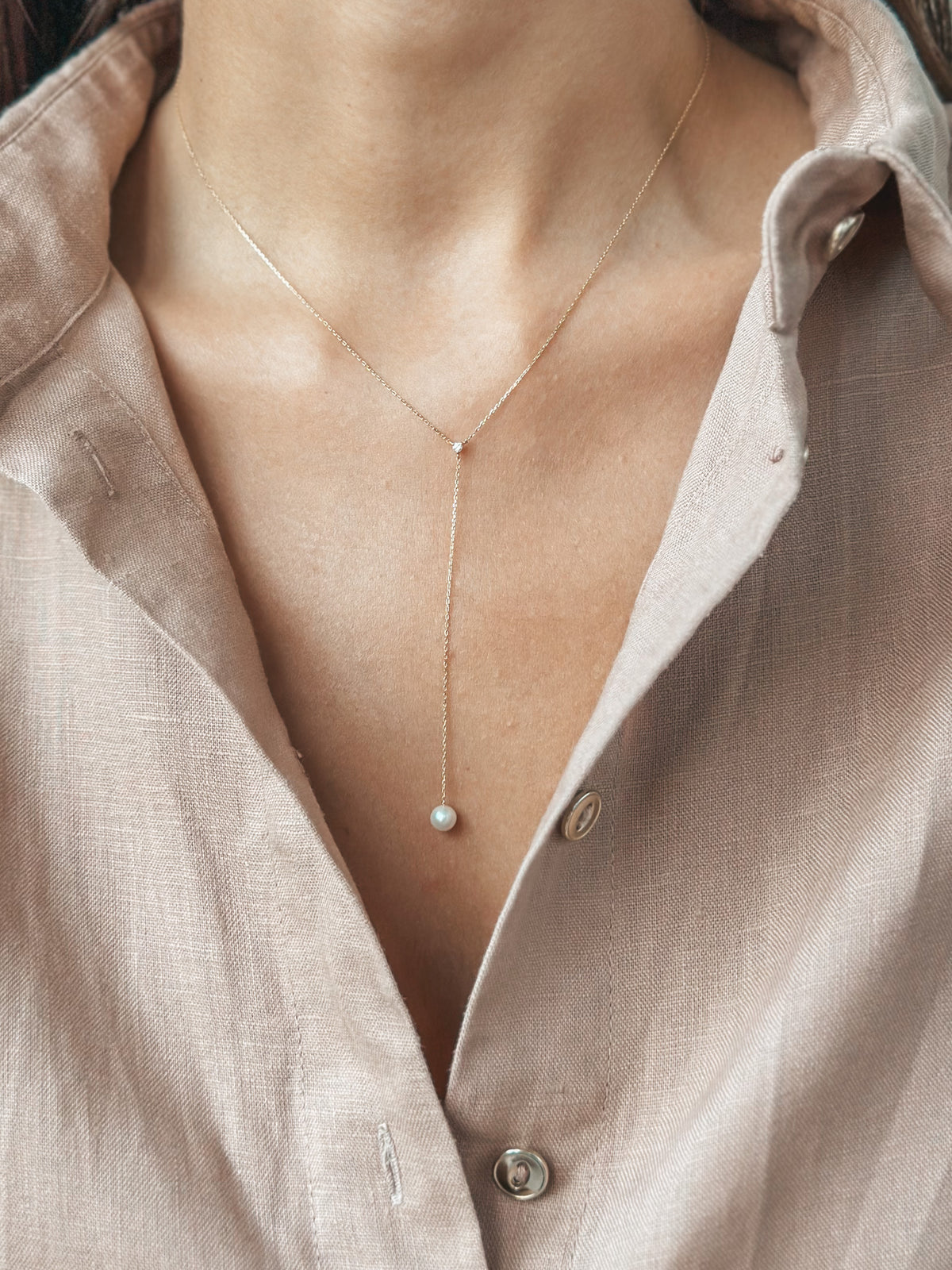 Close-up of a delicate gold necklace with a pearl pendant worn over an open-collared shirt.