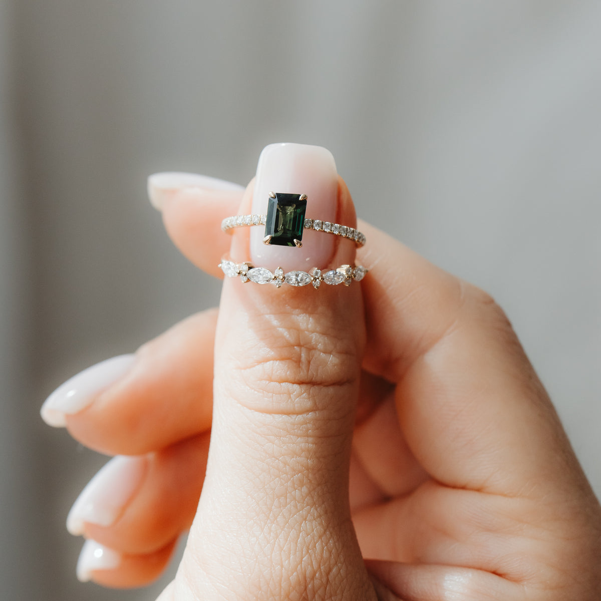 Nahaufnahme einer Hand, die zwei gelbgoldene Ringe hält: ein Elysian-Ring mit grünem Saphir im Emerald-Schliff und diamantbesetzter Pavé-Schiene, gestapelt über einem Fiorelle-Bandring mit Diamanten im Marquise-Schliff, vor softgrauem Hintergrund.