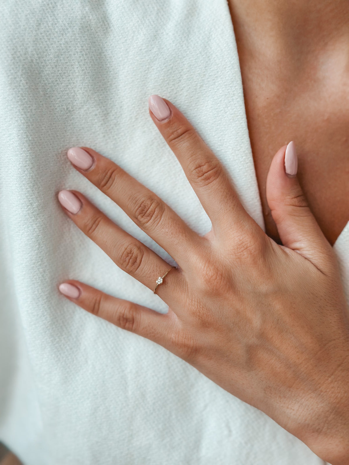 Nahaufnahme einer Hand mit rosa lackierten Nägeln, die einen filigranen Goldring mit einem kleinen Diamanten vor einem weichen weißen Stoffhintergrund trägt.