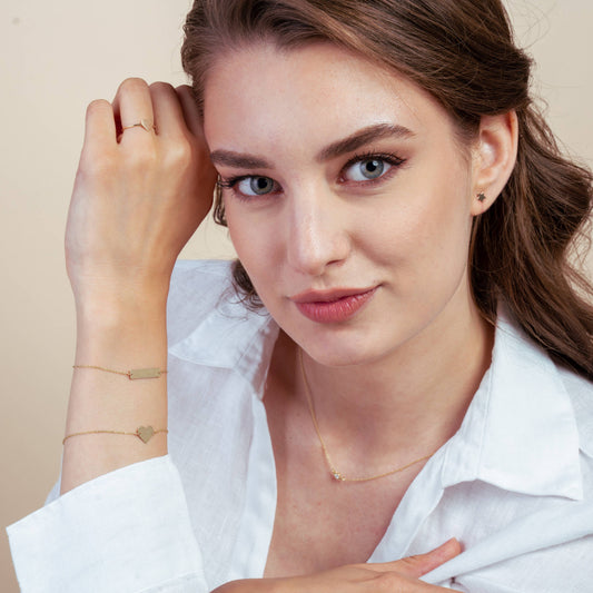 Frau in weißem Hemd präsentiert goldenen Schmuck – ein Herzarmband, kleine Ohrringe und eine zarte Halskette – im Studio.