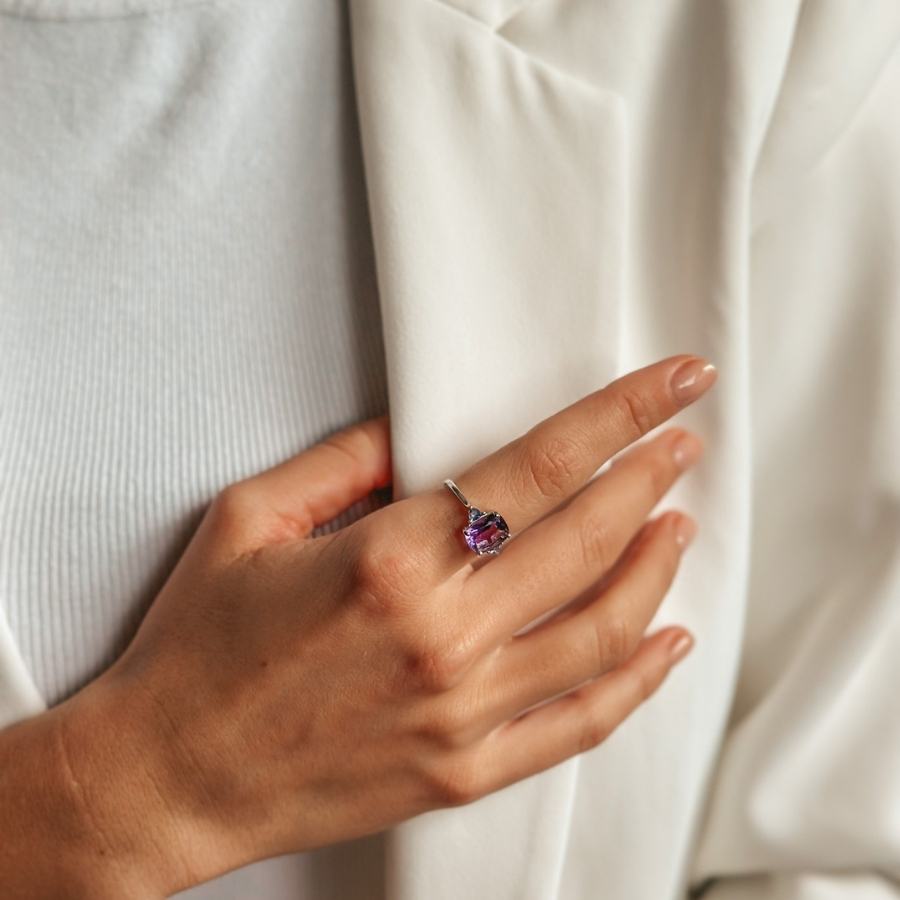 Nahaufnahme einer Hand, die einen Silberring mit Amethyst- und Tansanit-Edelsteinen trägt, ruhend auf einem weißen Kleidungsstück.